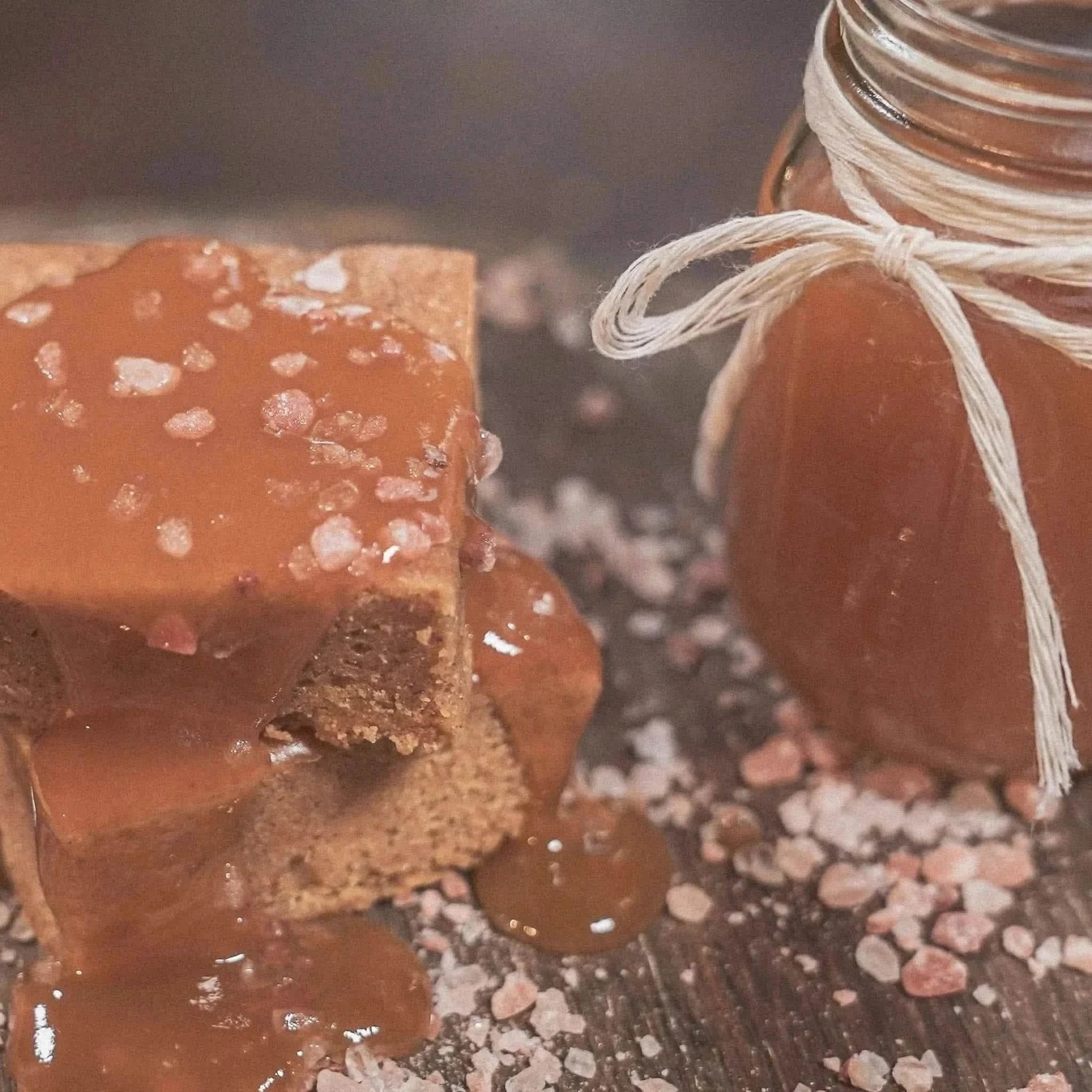 A salted caramel blondie topped with rich caramel sauce and pink Himalayan salt crystals, placed on a wooden surface next to a jar of homemade caramel tied with twine.