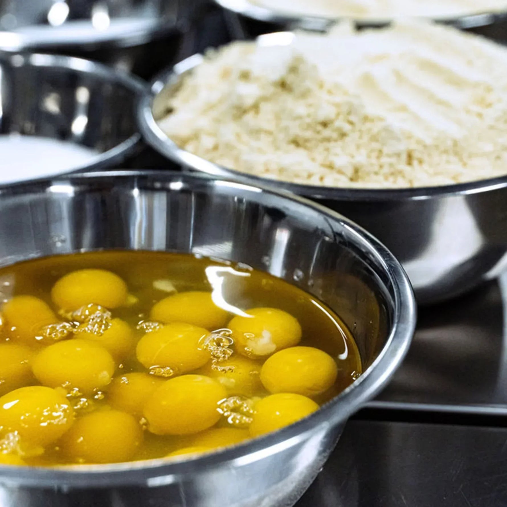 Stainless steel mixing bowls filled with fresh cracked eggs and almond flour, set on a clean commercial kitchen countertop, ready for baking.