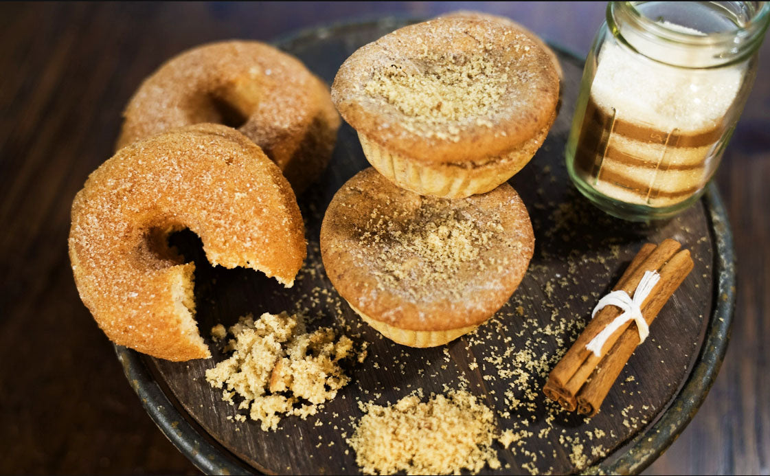 Close-up of cinnamon donuts and muffins on a rustic wooden platter, garnished with powdered erythritol and cinnamon, alongside a jar of erythritol and tied cinnamon sticks. Perfect for showcasing baked goods or cinnamon-flavored snacks.