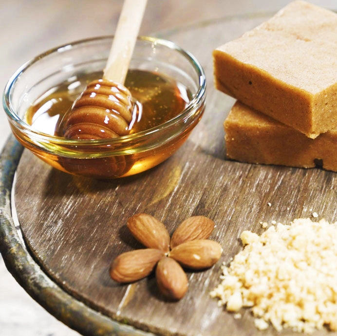 A wooden platter featuring a bowl of golden honey with a honey dipper, two almond-based snack bars, raw almonds, and ground almond flour, showcasing the natural and wholesome ingredients used in Protein Snack Shop products.
