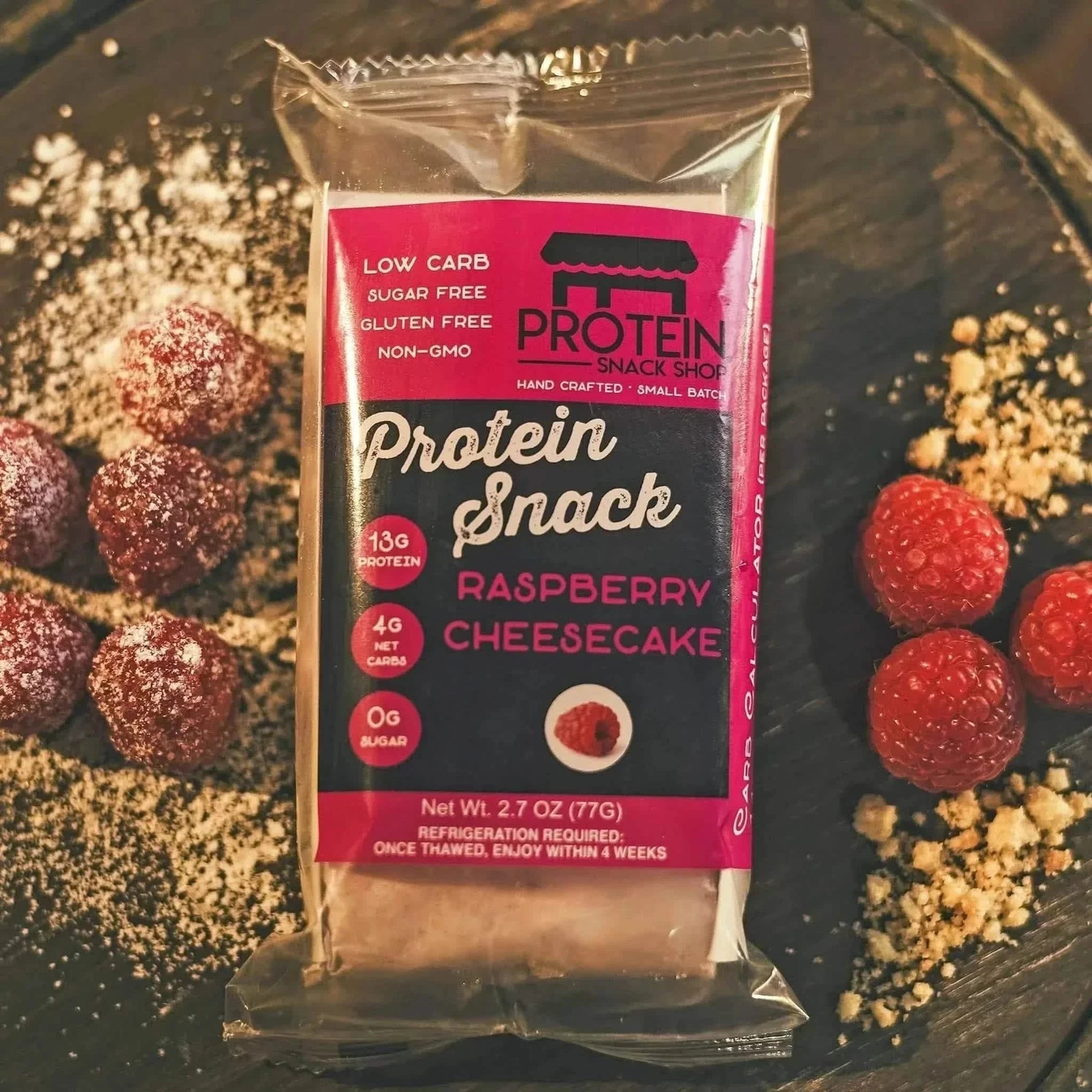 Packaged Raspberry Cheesecake Protein Snack from Protein Snack Shop, preserved with nitrogen packaging to ensure freshness. Displayed on a rustic wooden surface alongside fresh raspberries, powdered erythritol.
