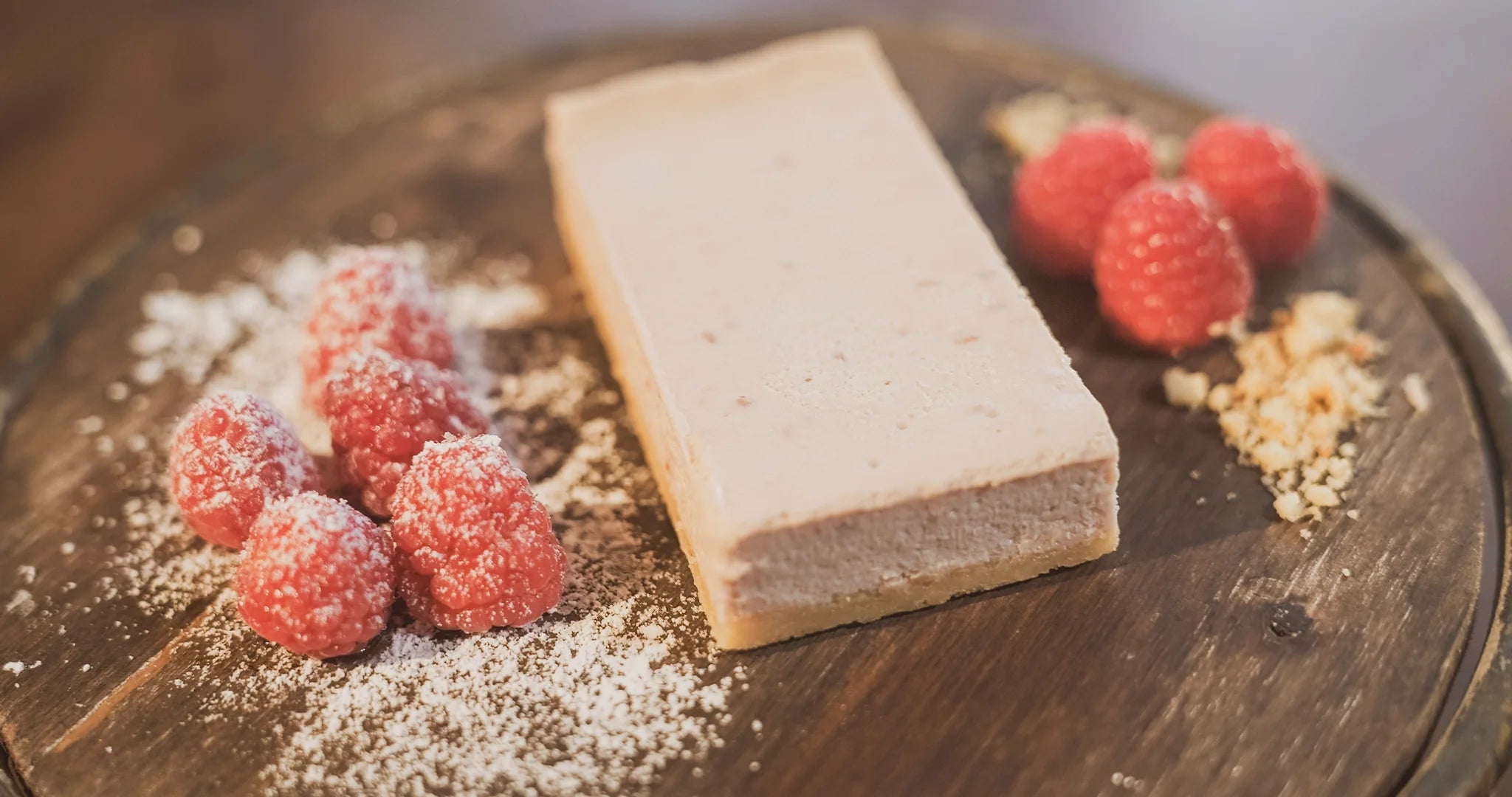 A decadent slice of raspberry protein cheesecake on a wooden plate, garnished with fresh raspberries lightly dusted with powdered erythritol.