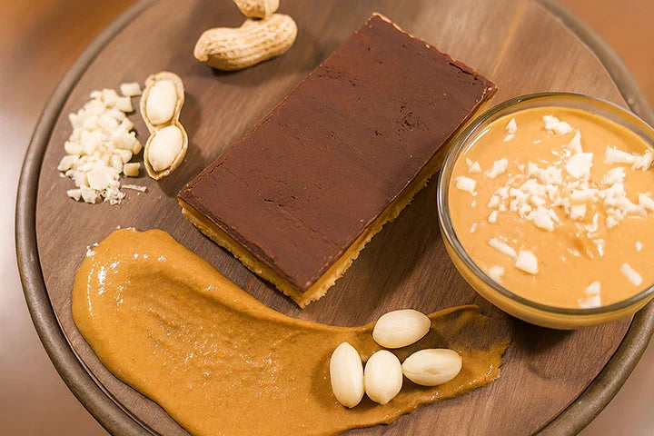 Peanut butter chocolate bar displayed on a wooden board with a bowl of creamy peanut butter, whole peanuts, chopped peanuts, and a peanut butter smear.