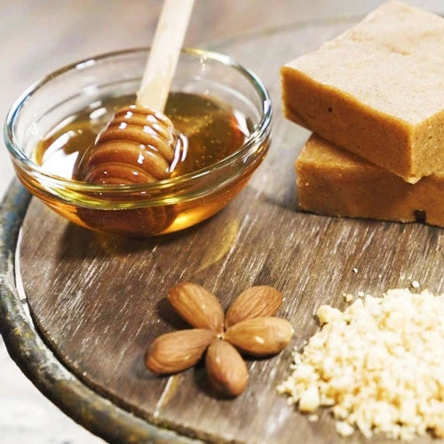 Honey in a glass bowl with a wooden dipper, almonds, and clean-ingredient blondie bars on a rustic wooden board.
