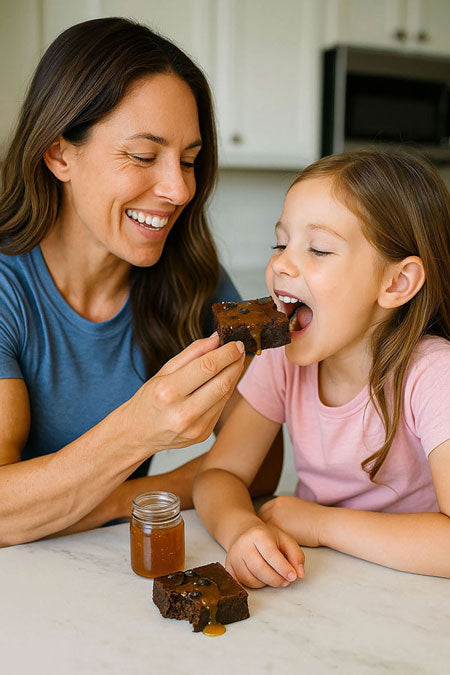 Mother smiling as she shares a clean-ingredient chocolate protein brownie with her young daughter at the kitchen table.
